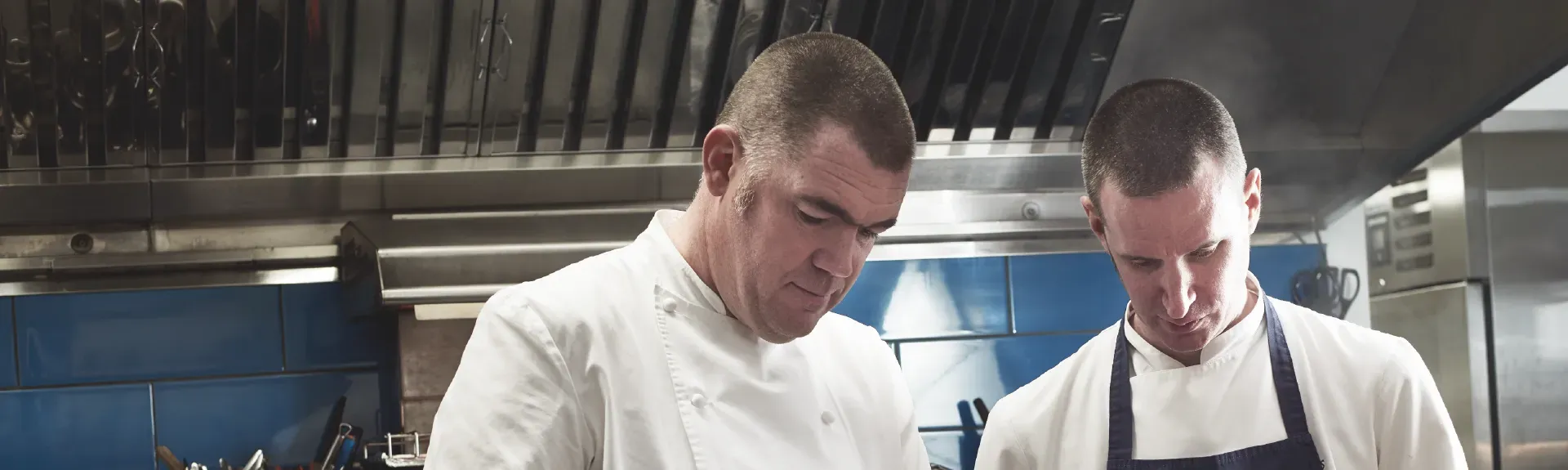 Two male chefs in white uniforms focus on preparing meat in a professional kitchen.